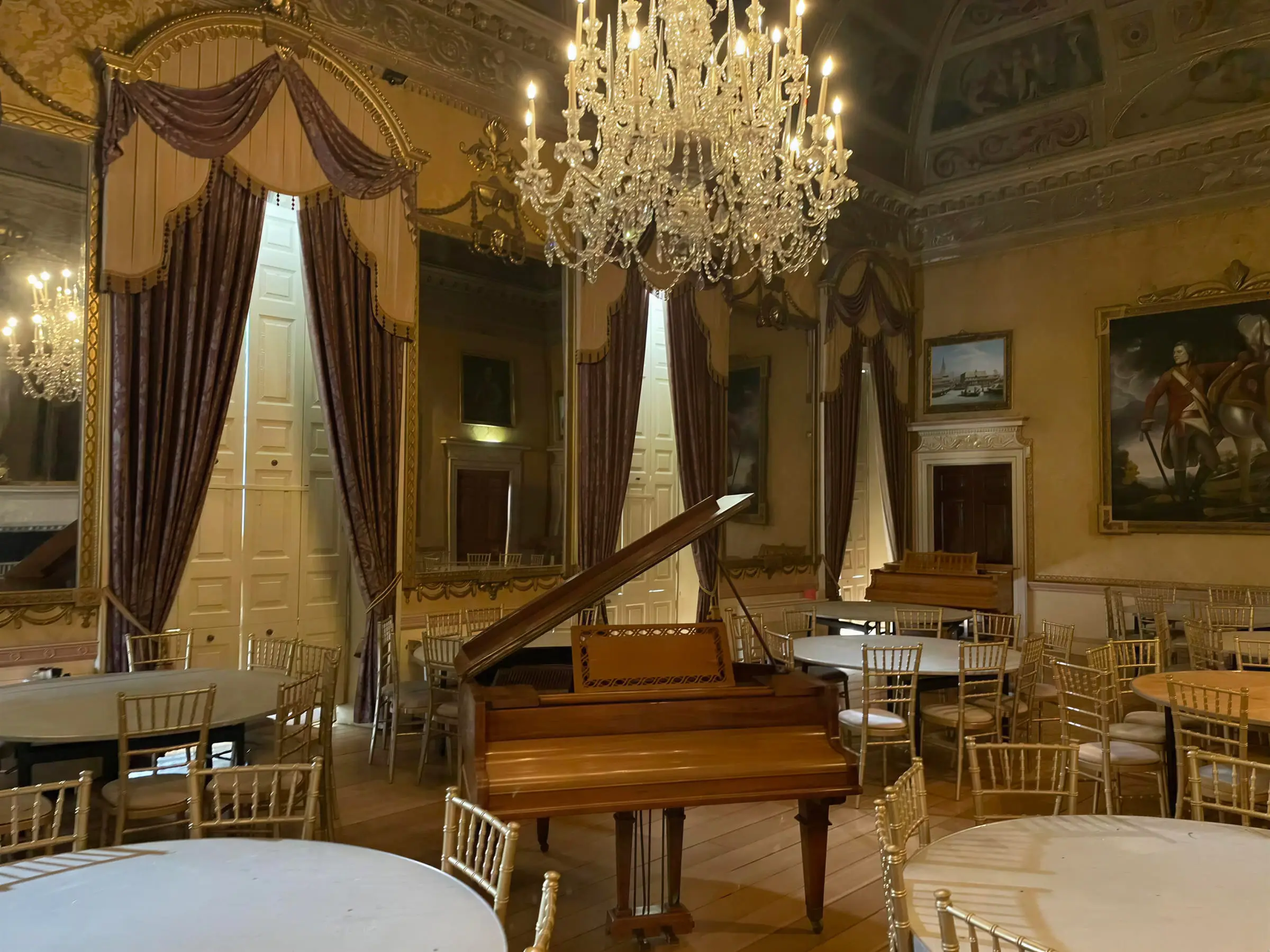 A baby grand piano standing open in a gilded ballroom beneath a chandelier