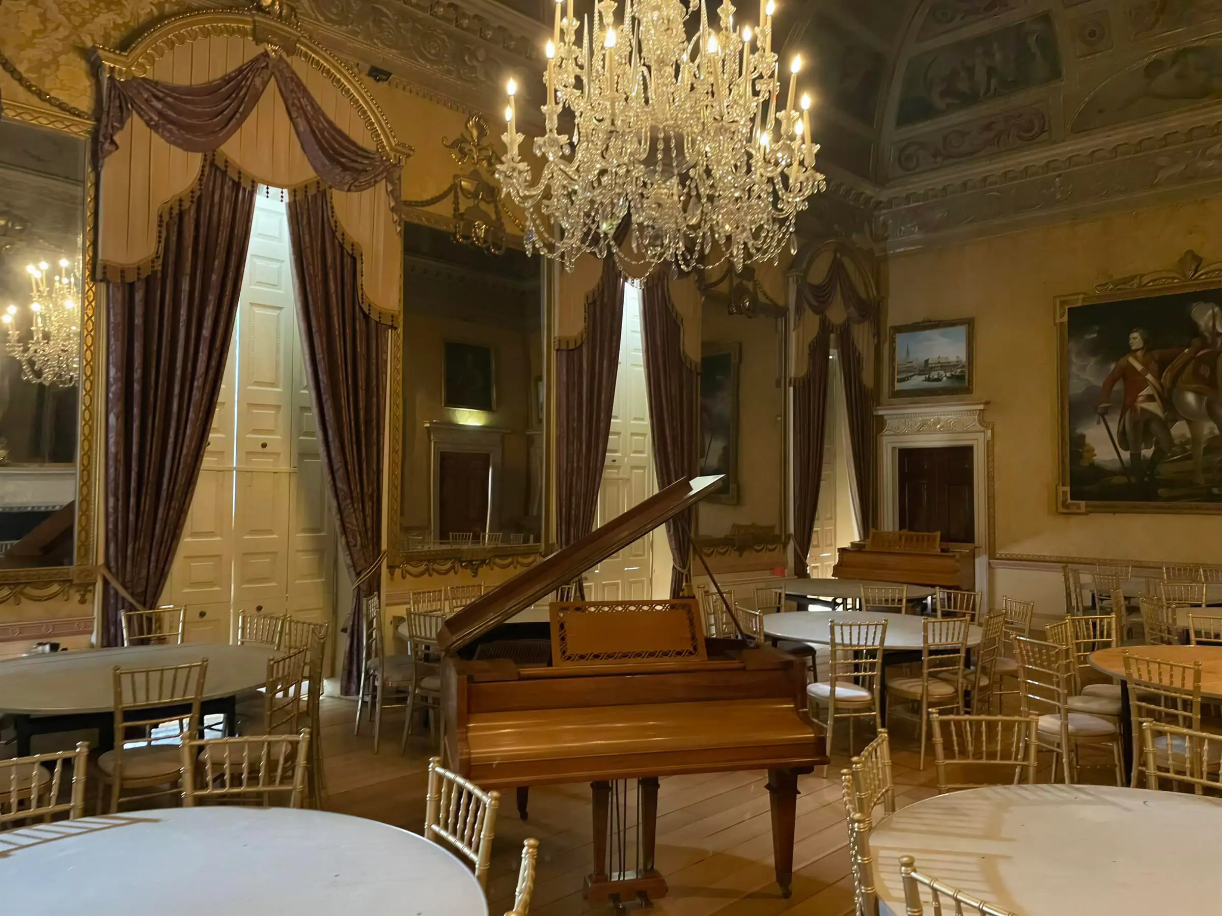A baby grand piano standing open in a gilded ballroom beneath a chandelier
