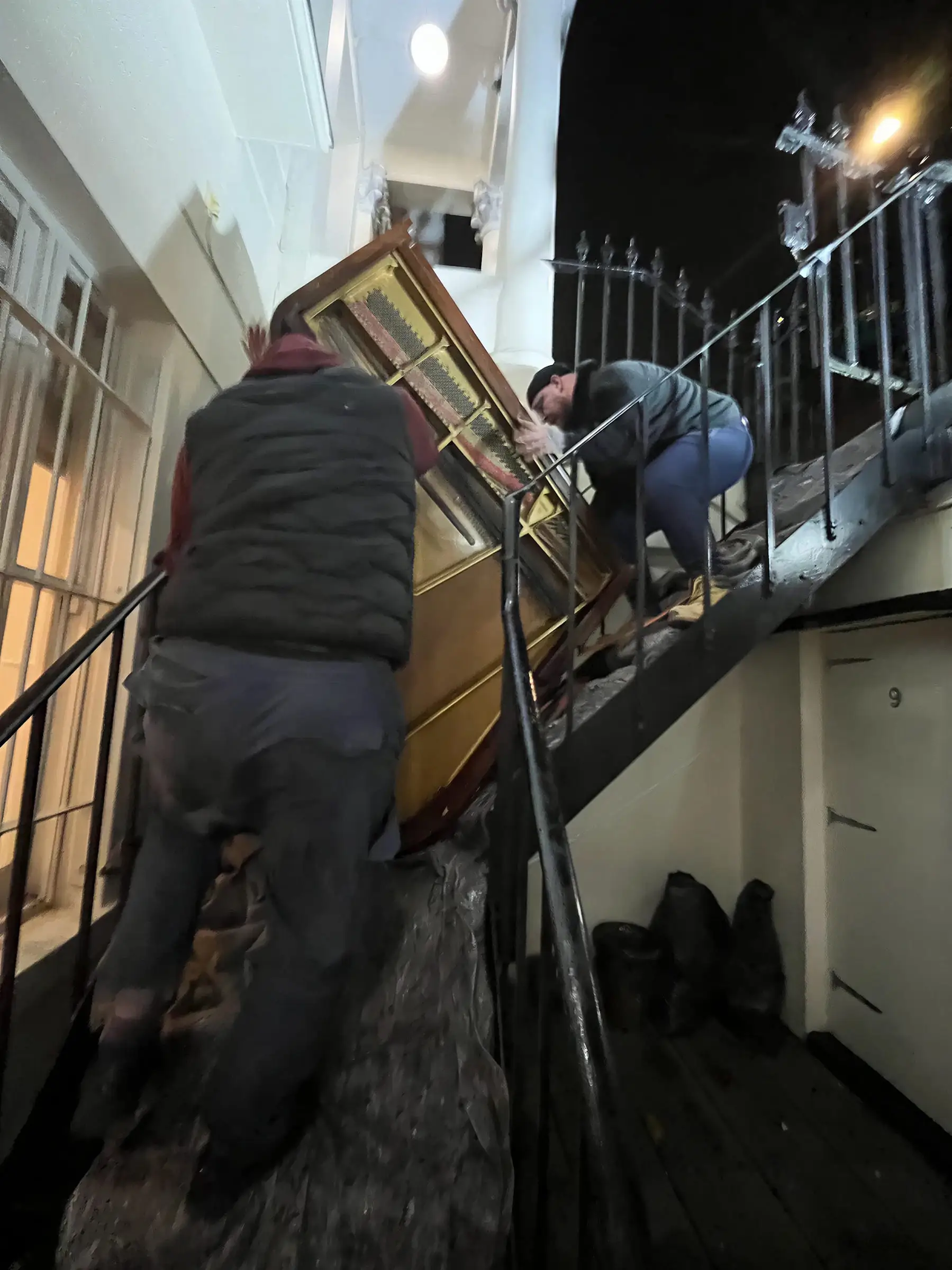 Two movers carrying a baby grand piano on its side down a narrow unlit staircase