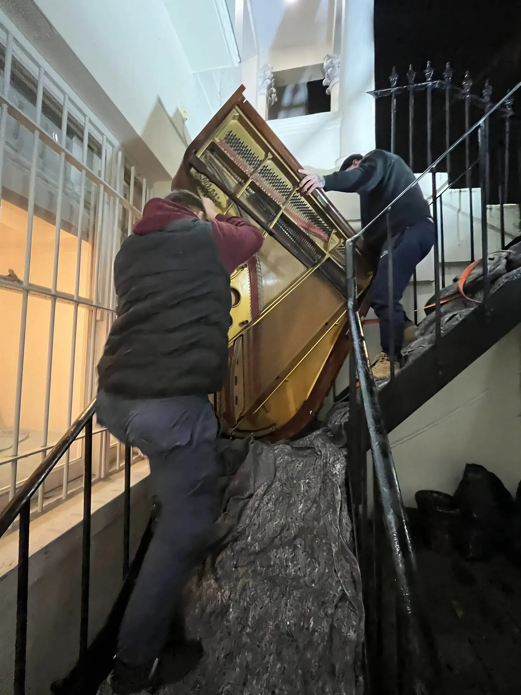 A baby grand piano on its side on a narrow staircase, one mover below it taking the weight and one above
