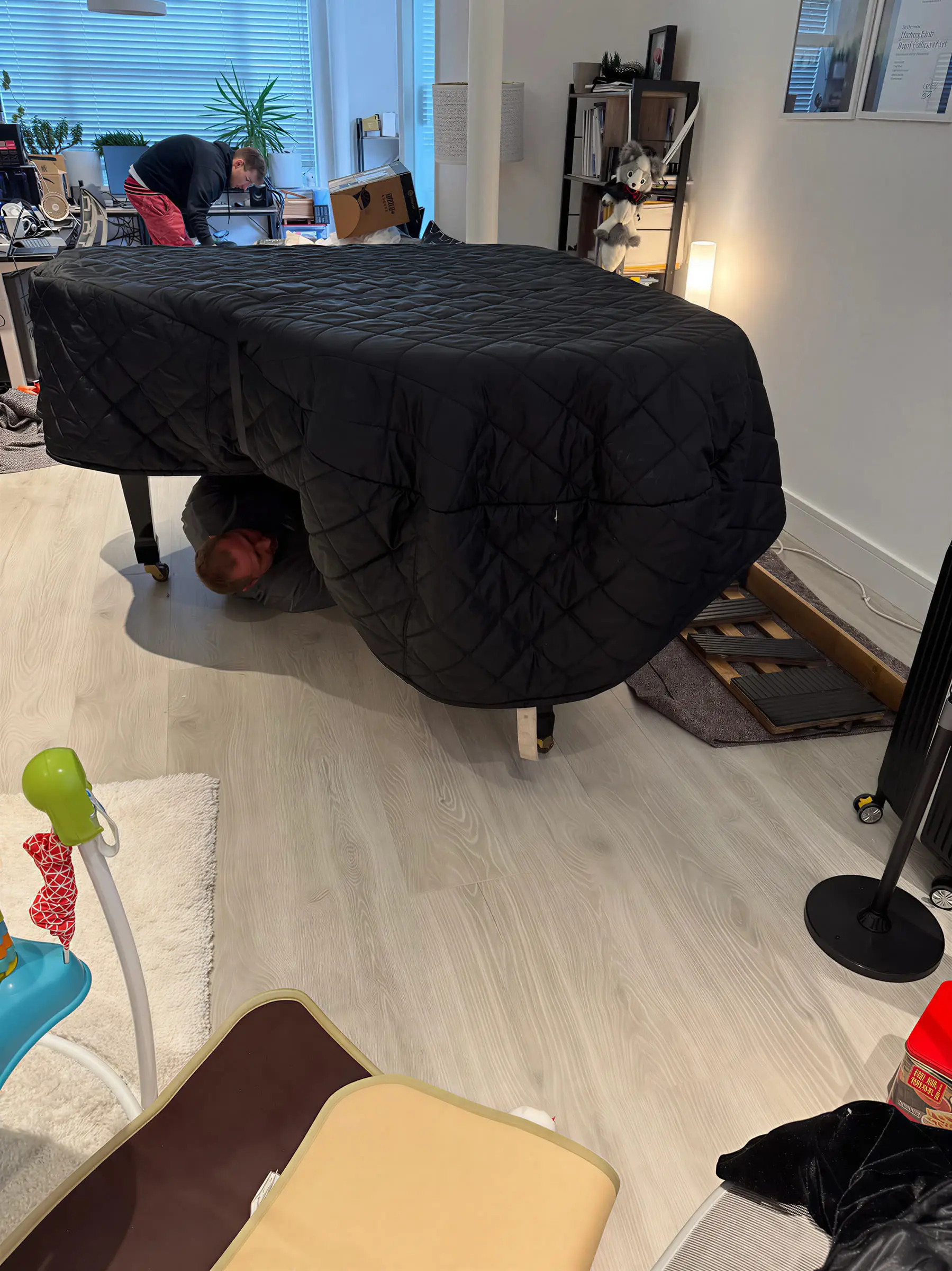A grand piano wrapped in a quilted cover in a lounge, with a mover working underneath it