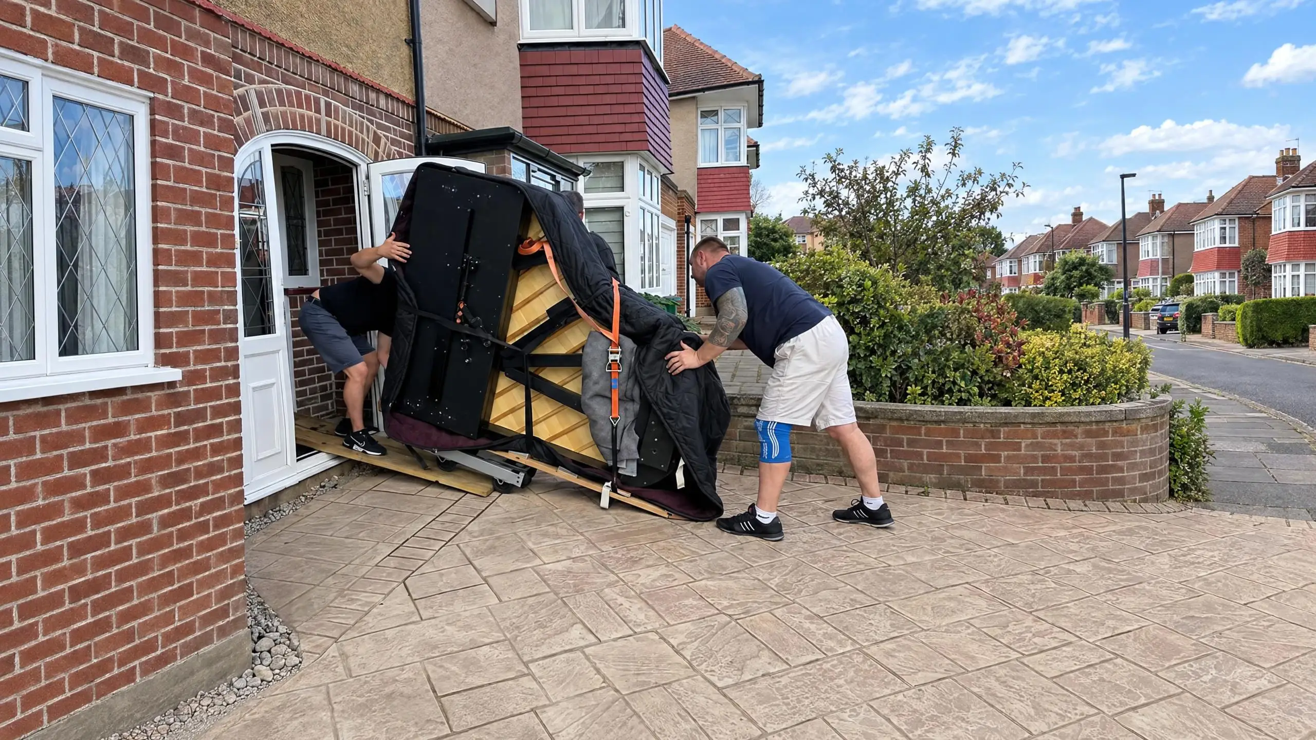 Two VVM movers easing a grand piano on its side in through the front door of a house