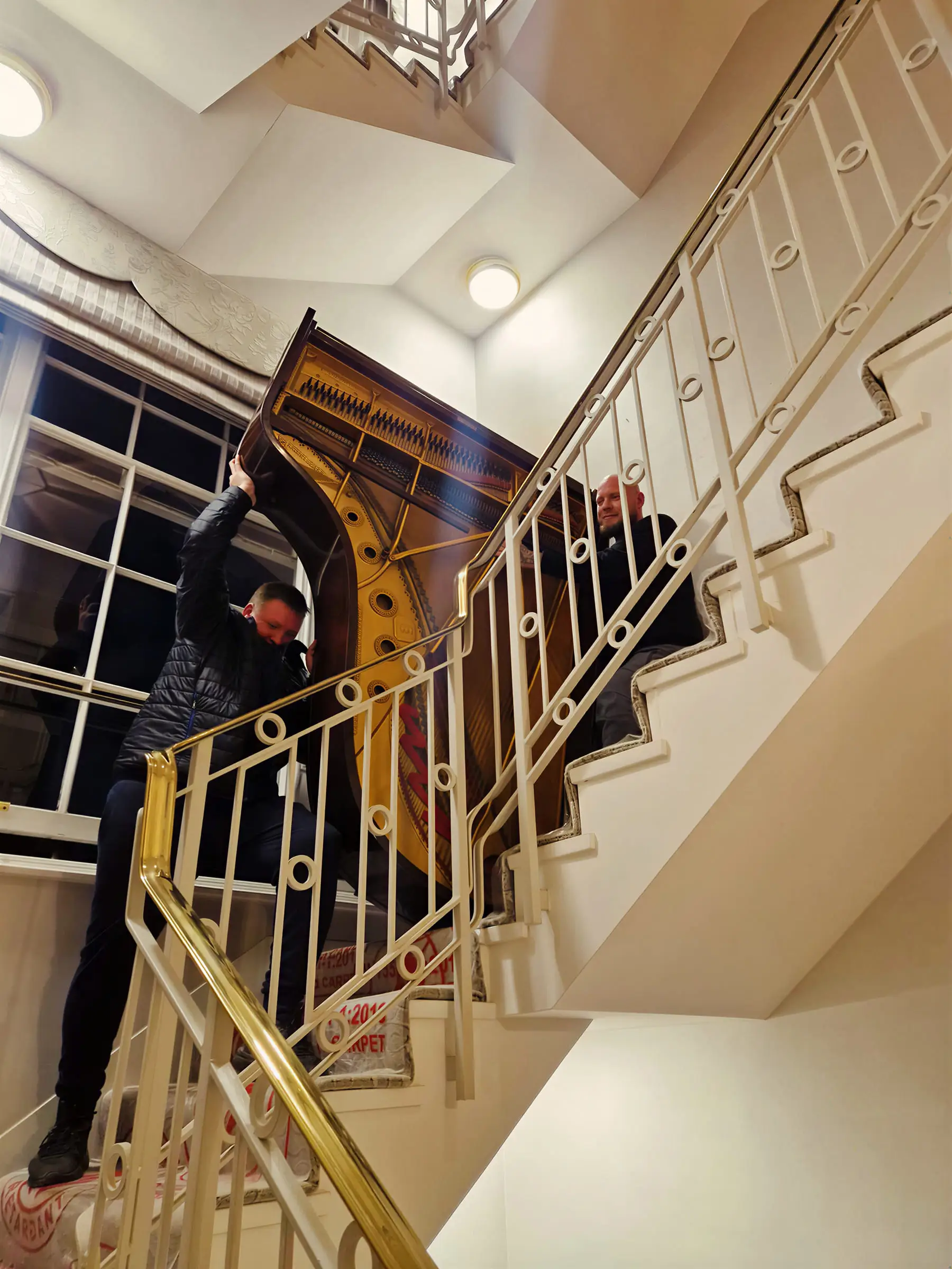A piano being carried up a bright stairwell past a brass handrail