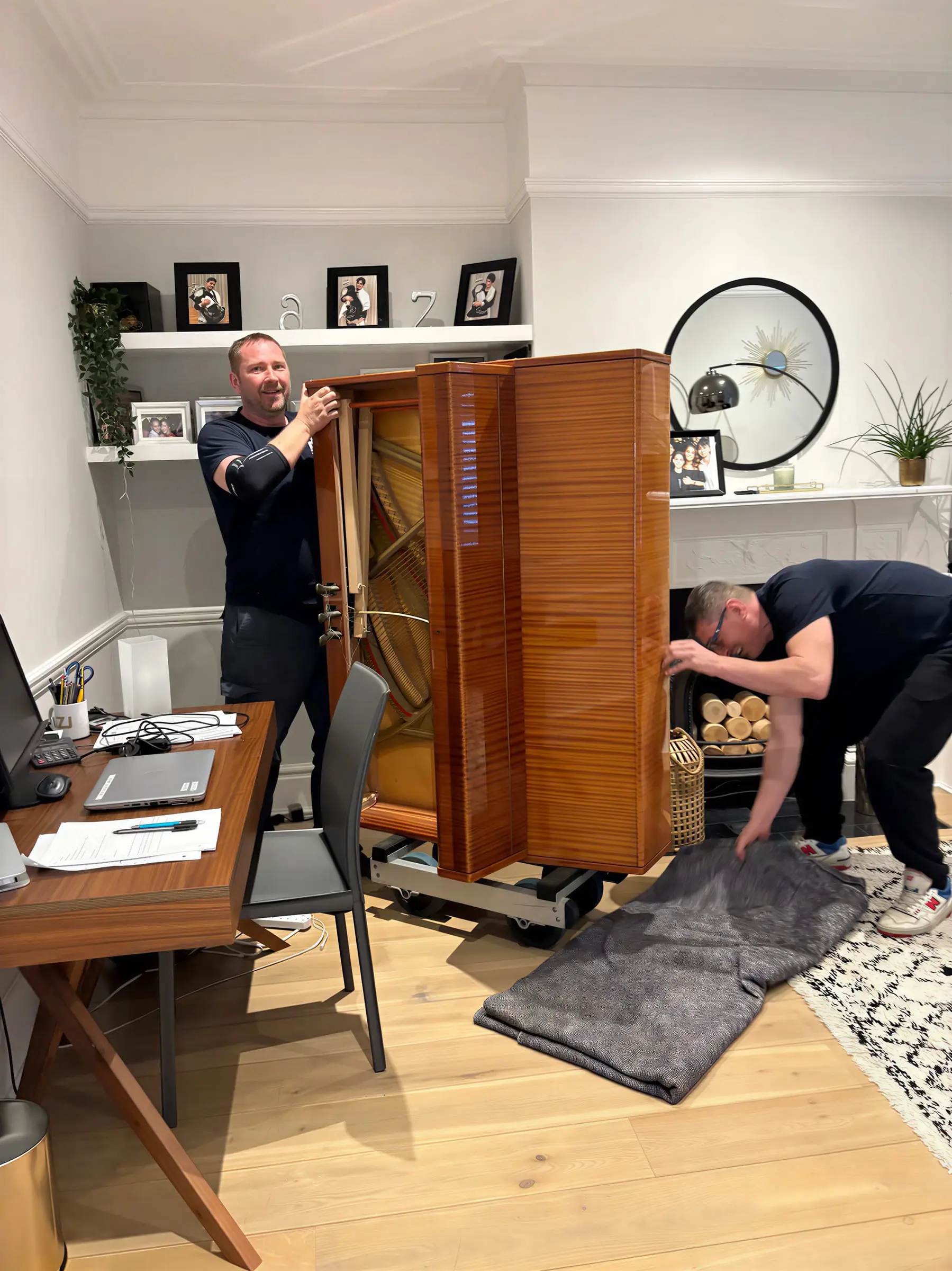 Two movers setting down a tall wooden upright piano in a home office