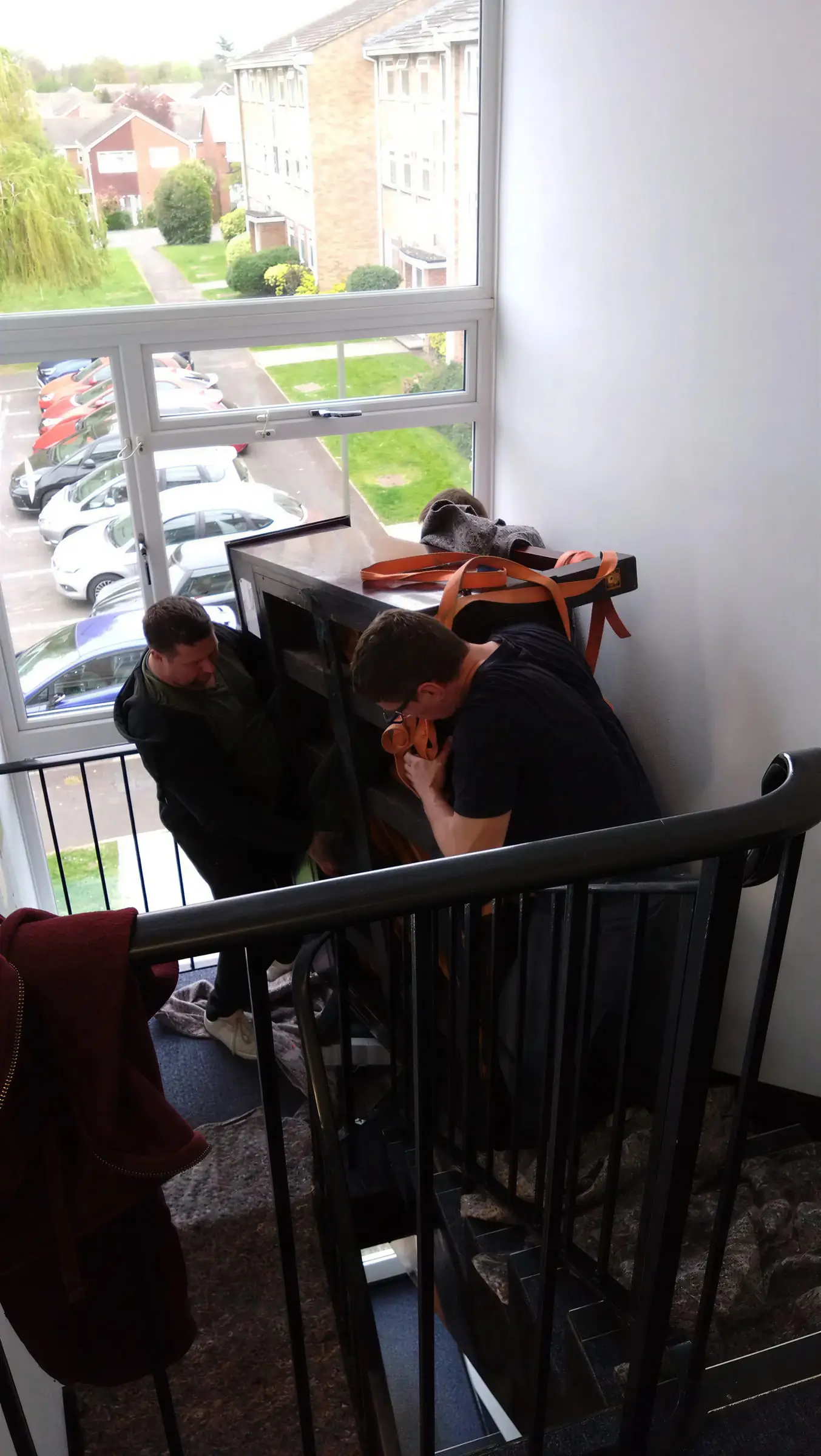 Two movers turning the same upright piano on the landing beside a window