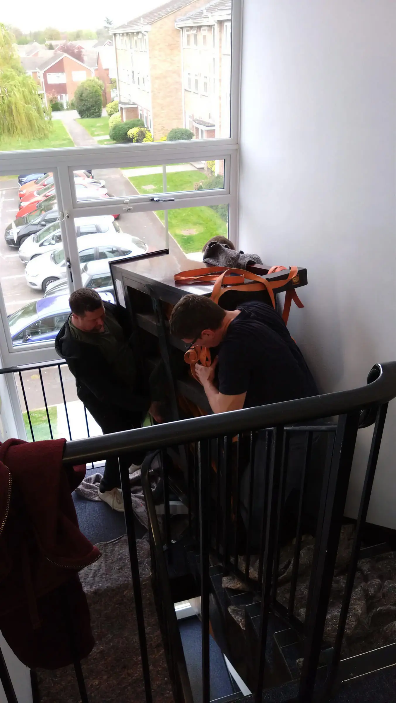 Two movers turning the same upright piano on the landing beside a window